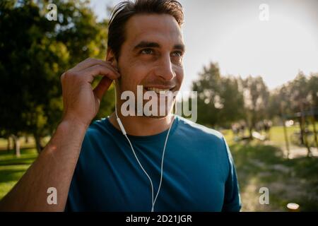 Male young athlete adjusting ear phones to listen to music smiling and resting from exercising outdoors in sun Stock Photo
