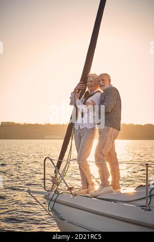 Full leghth of beautiful happy senior couple in love standing on the side of sail boat floating in sea at sunset, they are hugging and enjoying Stock Photo