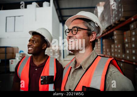 caucasian male worker wearing spectacles laughing with mixed race worker standing in factory warehouse. Stock Photo