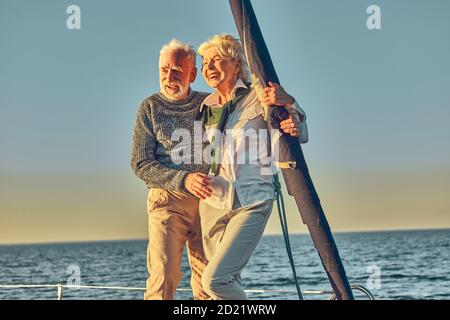 Happy senior couple in love, retired man and woman standing on the side of sail boat or yacht deck floating in sea, enjoying amazing sunset and Stock Photo