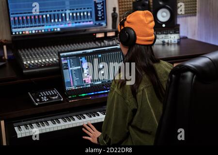 Young woman, female artist looking focused while playing keyboard synthesizer, sitting in recording studio Stock Photo