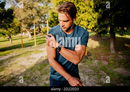Injured young male athlete looking concerned holding shoulder in pain after exercising outdoors with smartwatch and ear phones Stock Photo