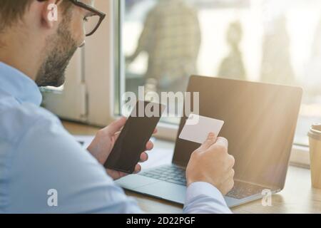 Important call. Rear view of a young businessman or male freelancer holding smartphone and looking at blank business card while working remotely in Stock Photo
