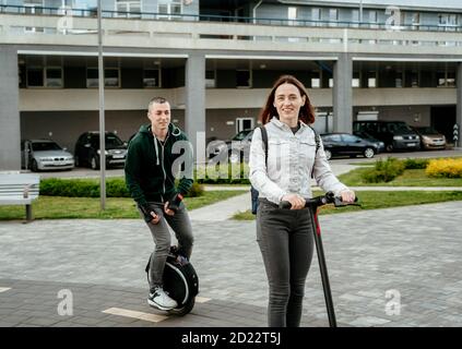 Young man riding unicycle and young woman in casual wear riding on electric kick scooter on city street Stock Photo