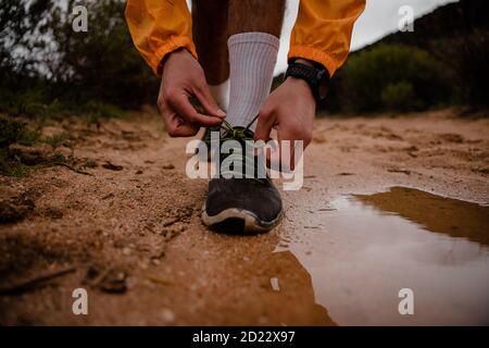 Adventurous young male athlete tying shoe laces while running on wet mountain gravel path with luscious bushes Stock Photo