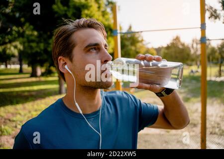 Thirsty young male athlete drinking water looking upwards while listening to music in ear phones exercising outdoors Stock Photo