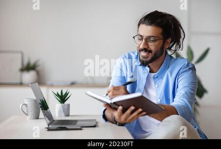 Smiling Western Freelancer Guy Taking Notes At Workplace In Home Office Stock Photo
