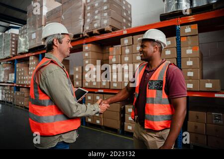 Two male colleagues shaking hands in congratulation of successful business deal in packaging department standing in warehouse Stock Photo