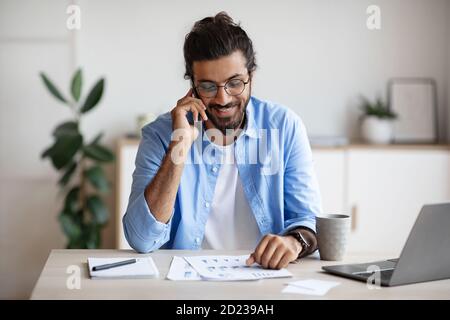 Freelancer Guy Checking Papers And Talking On Cellphone, Working At Home Office Stock Photo