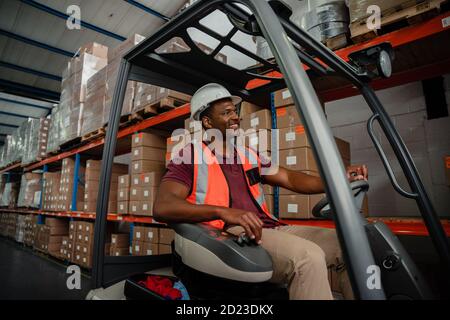 Mixed race worker driving forklift around warehouse while searching for parcels  Stock Photo