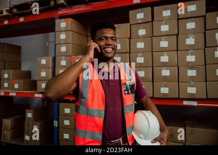 Male worker holding hardhat chatting to colleagues on cellular smartphone standing next to parcels. Stock Photo