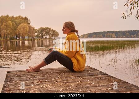 A barefoot girl with her hair in braids sits on a wooden bridge by the lake against the sunset. The girl is holding a Cup with a drink.  Stock Photo