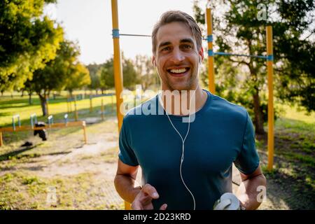 Handsome young male athlete listening to music in ear phones before training on the outdoor equipment in the sunny park Stock Photo