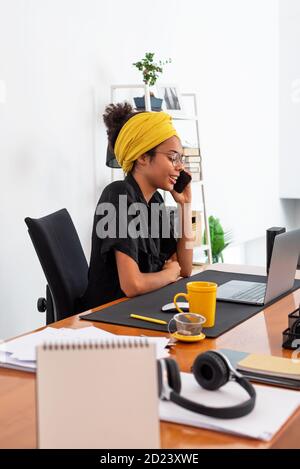 Shot of young latin woman working at home office with laptop speaking on mobile phone Stock Photo
