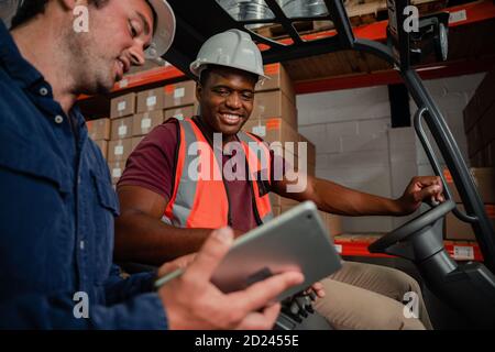 Caucasian worker holding digital tablet showing ethnic worker location of parcel driving forklift in warehouse Stock Photo