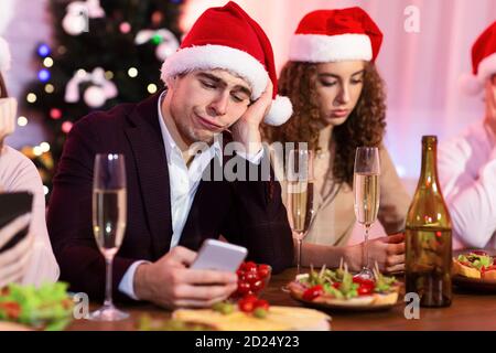 Bored Guy Using Smartphone During Christmas Party With Friends Indoor Stock Photo