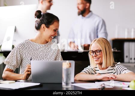 Two laughing businesswomen talking together over a laptop at an office desk with colleagues working in the background Stock Photo