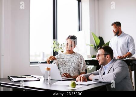 Two smiling businesspeople discussing work on a laptop at an office desk with a colleague working in the background Stock Photo