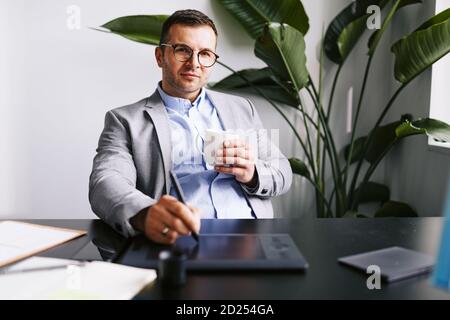Businessman sitting at his desk drinking coffee and working on a computer using a graphics tablet Stock Photo