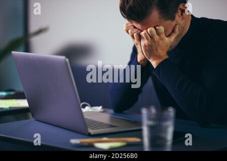 Stressed-out young businessman sitting with his head in his hands while working on a laptop at an office desk Stock Photo