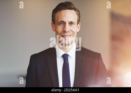 Confident young businessman in a suit smiling while standing alone in a bright modern office Stock Photo