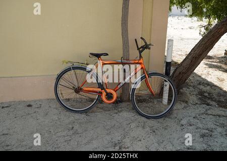 An old orange bicycle is standing on the sand path locked to a tree. Old Apache orange cycle chained to a tree. Man's Cycle with lock in tree in a san Stock Photo