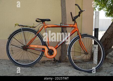 An old orange bicycle is standing on the sand path locked to a tree. Old Apache orange cycle chained to a tree. Man's Cycle with lock in tree in a san Stock Photo