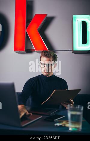 Young businessman sitting at a desk in a modern office going over paperwork on a clipboard and using a laptop Stock Photo