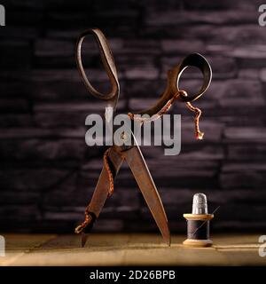 Rusty vintage pair of scissors balanced on timber flooring with black cotton reel thread needle and silver thimble against slate background Stock Photo