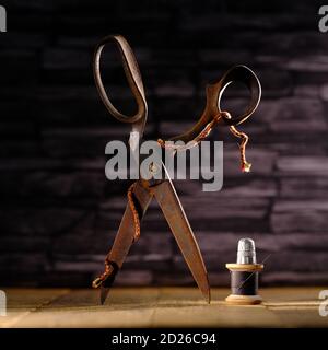 Rusty vintage pair of scissors balanced on timber flooring with black cotton reel thread needle and silver thimble against slate background Stock Photo