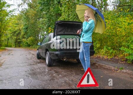 A slender blonde girl stands near a broken car with an umbrella in her hands and talks on the phone and asks for help Stock Photo