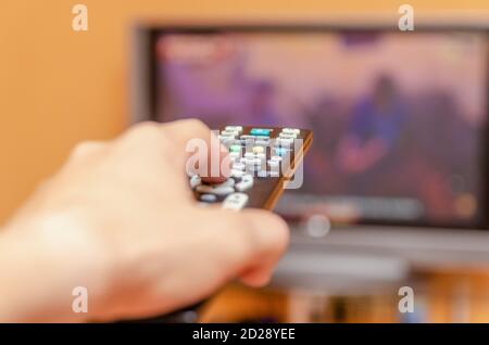 Man's hand holding a tv remote control, pressing a button while pointing at a flat screen tv. Stock Photo