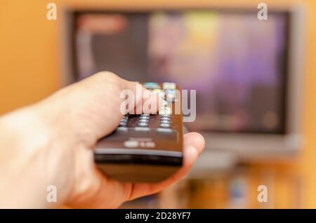 Man's hand holding a tv remote control, pressing a button while pointing at a flat screen tv. Stock Photo