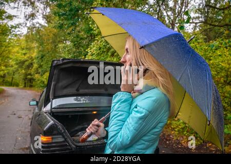 A slender blonde girl stands near a broken car with an umbrella in her hands and talks on the phone and asks for help Stock Photo