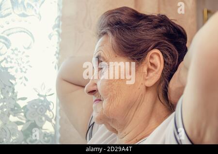 elderly dark haired woman with her hands behind her head looks out the window Stock Photo