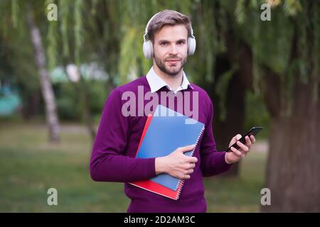 check his email. man listen music in headphones and hold notebook. handsome guy in earphones relax in park use phone. modern life education technology. student get information from book. study online. Stock Photo