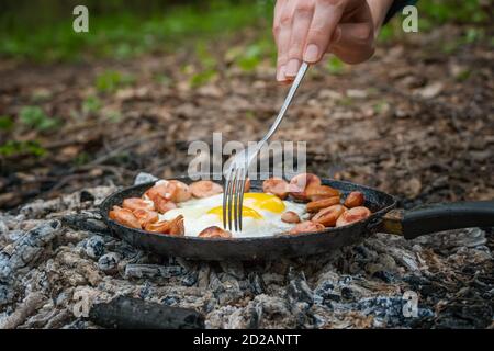 Girl's hand with a fork turns over pieces of fried sausage and eggs in a pan on the coals. Breakfast in nature, picnic, camping food. Stock Photo