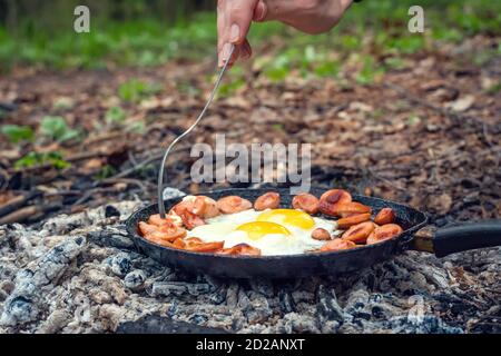 Girl's hand with a fork turns over pieces of fried sausage and eggs in a pan on the coals. Breakfast in nature, picnic, camping food. Stock Photo