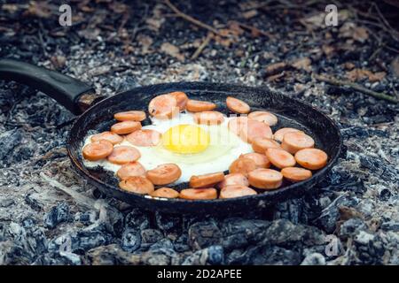 egg and sausages are fried in a pan on coals in the forest. Breakfast in the nature, picnic. Stock Photo