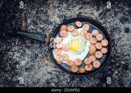 egg and sausages are fried in a pan on coals in the forest. Breakfast in the nature, picnic, view top Stock Photo
