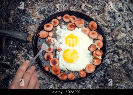 Girl's hand with a fork turns over pieces of fried sausage and eggs in a pan on the coals, Breakfast in nature, picnic, camping food. Stock Photo