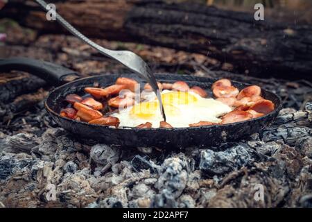 Girl's hand with a fork turns over pieces of fried sausage and eggs in a pan on the coals. Breakfast in nature, picnic, camping food. Stock Photo