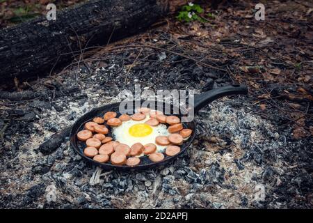 egg and sausages are fried in a pan on coals in the forest. Breakfast in the nature, picnic. Stock Photo