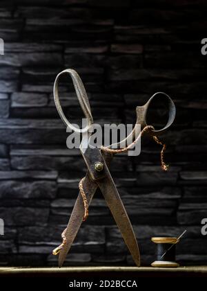 Rusty vintage pair of scissors balanced on timber flooring with black cotton reel thread needle and silver thimble against slate background Stock Photo