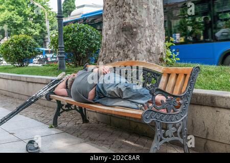 homeless man with a big belly sleeping on a wooden bench on a sunny day, next to crutches Stock Photo