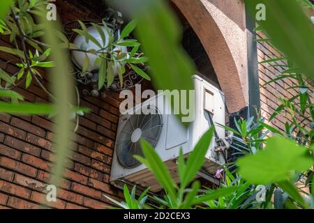 White air conditioner on a brick wall of a building outside among the leaves of trees Stock Photo