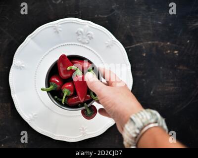 Female hand holding bowl of chilli peppers on white plate and dark table. Overhead shot with copy space. Stock Photo