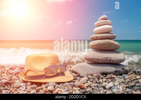 A cairn of stones near the sea, next to a straw hat and glasses. Stone tower on the beach on a sunny day. Balance, peace of mind. photo with backlight Stock Photo