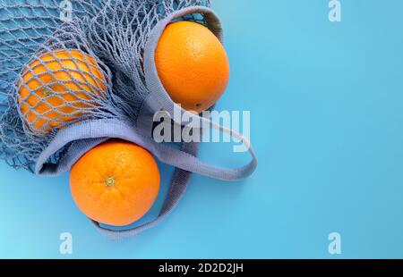 Three ripe oranges lie in a gray-blue eco-friendly string bag on a blue background. Stock Photo