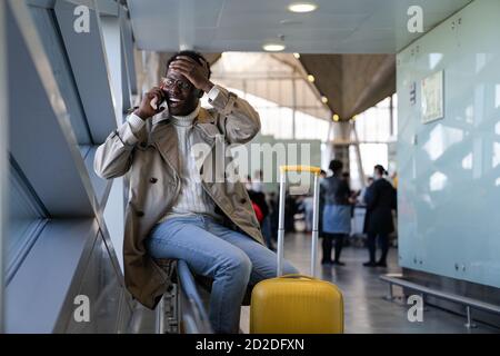 Stressed Black man in spectacles shocked with bad news, talking on mobile phone, sitting in airport, makes facepalm by hand and forgot something impor Stock Photo